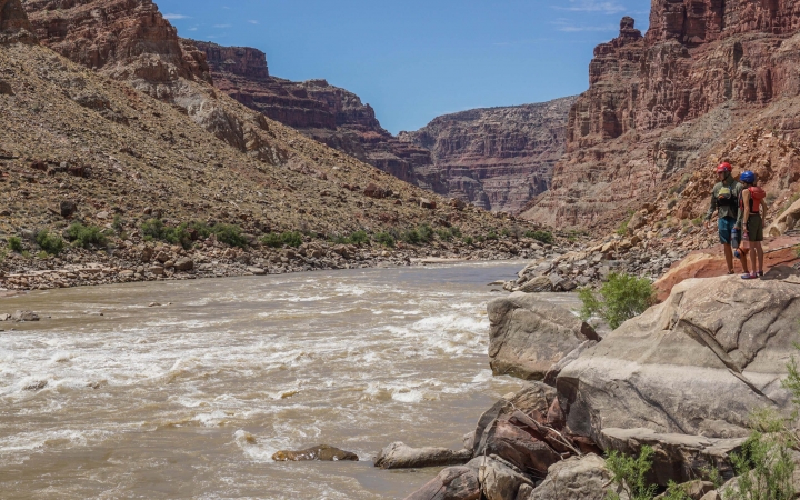 Two people stand on a rock overlooking a river below. There are tall canyon walls on either side of the river.